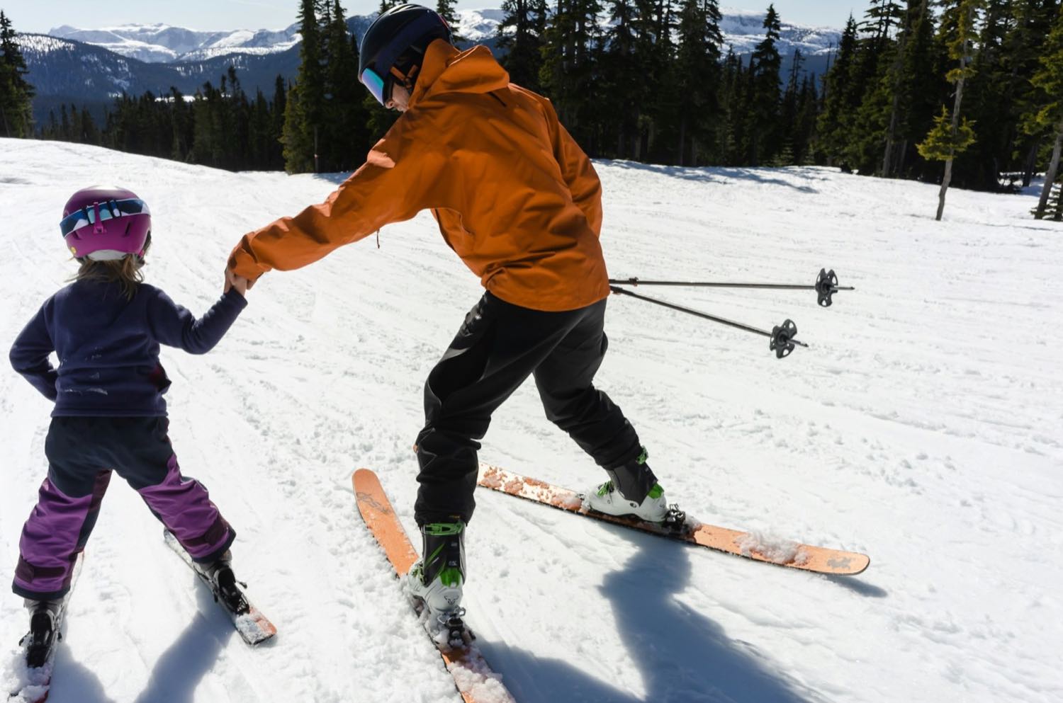 An adult in a bright jacket holding a small child's hand as she learns to ski on a gentle slope